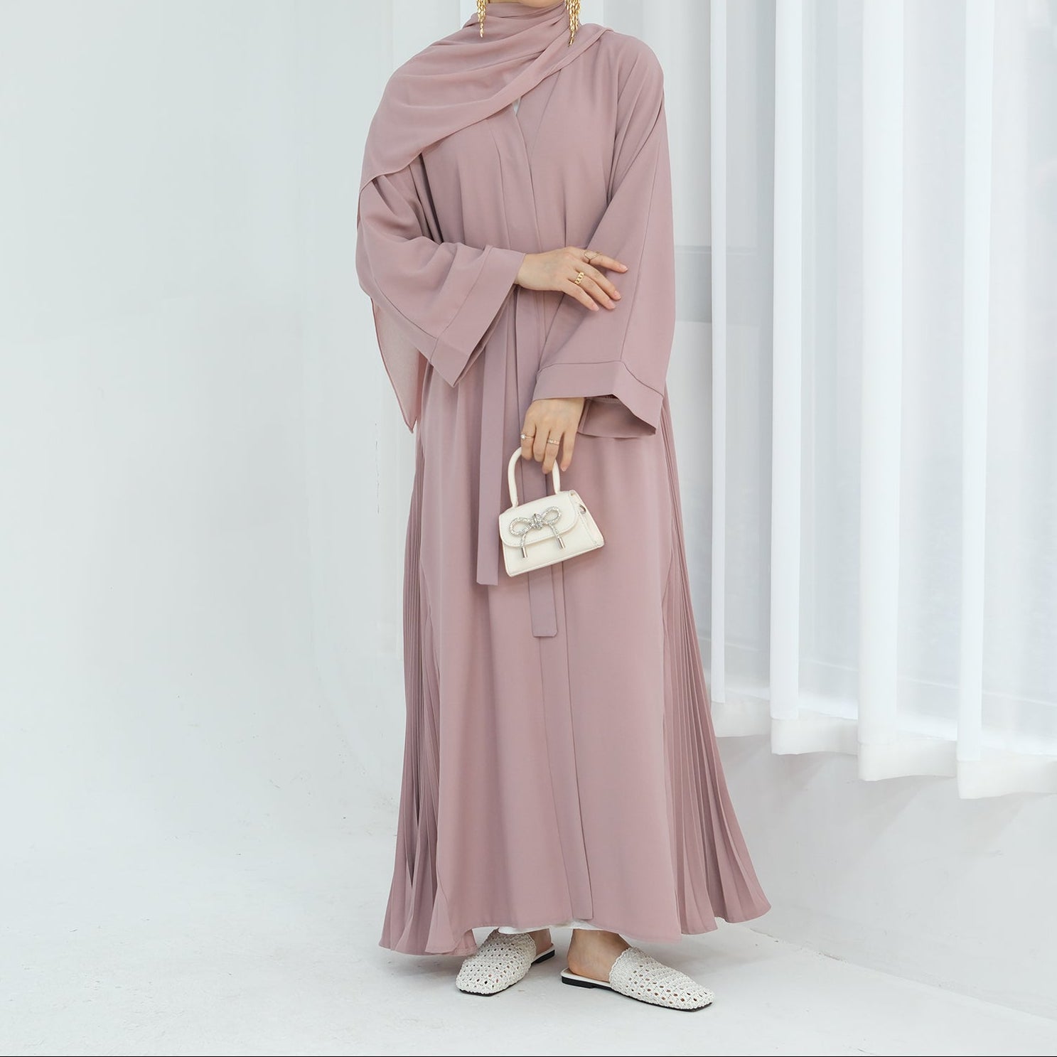 Woman wearing a pink hijab and dress holding a small handbag against a white curtain background. Noor & Grace