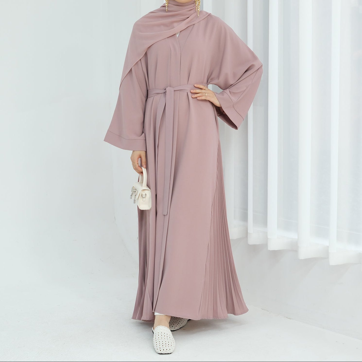 Woman wearing a long, light pink dress with a matching hijab against a white curtain background. Noor  & Grace