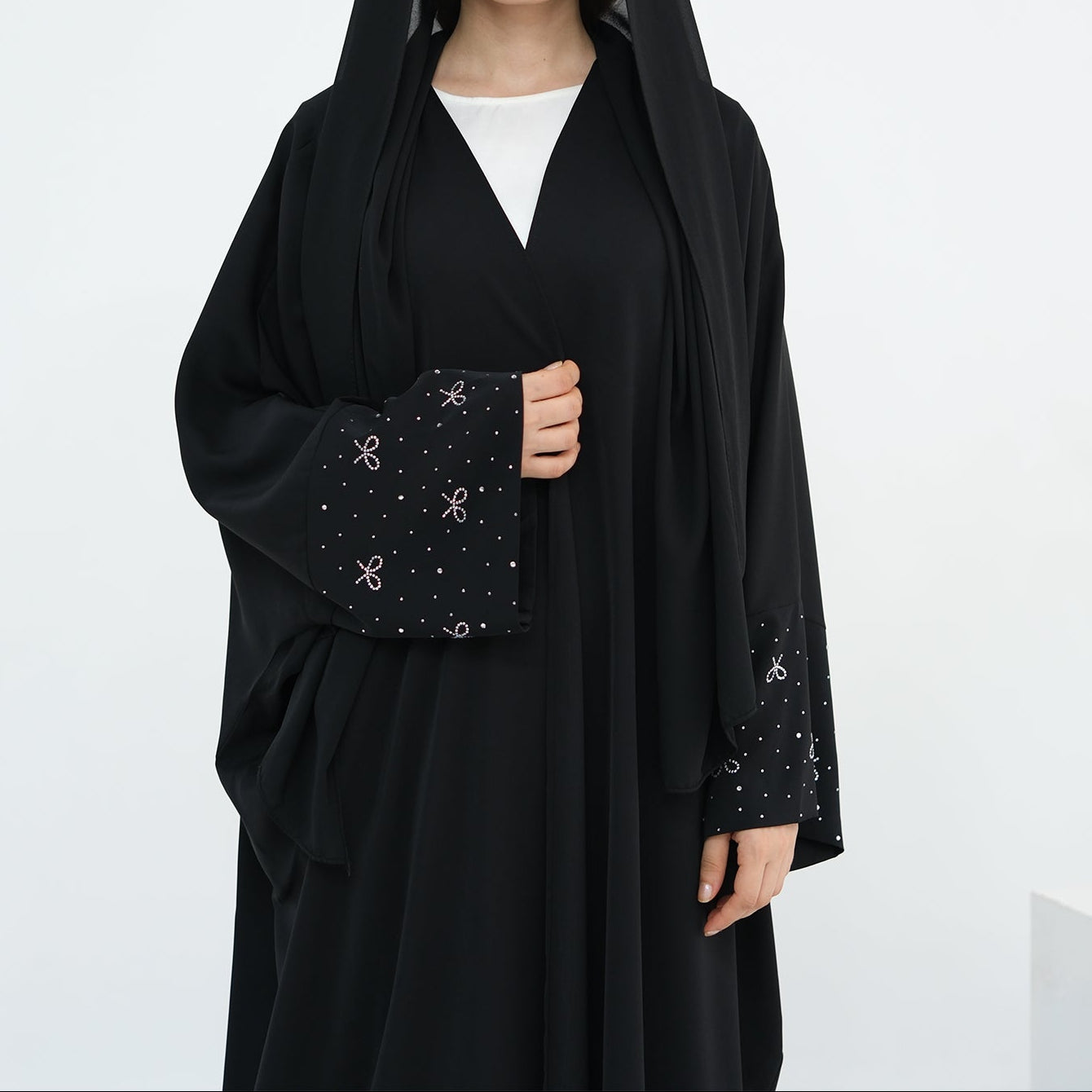 Woman wearing a black abaya with a white background. AlLayaliEmbroideredOpenAbaya_Noor_GraceBlack5
