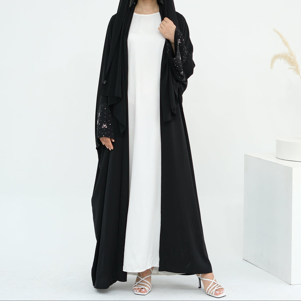 Woman wearing a black abaya over a white dress on a white background. AlLayaliEmbroideredOpenAbaya_Noor_GraceBlack8