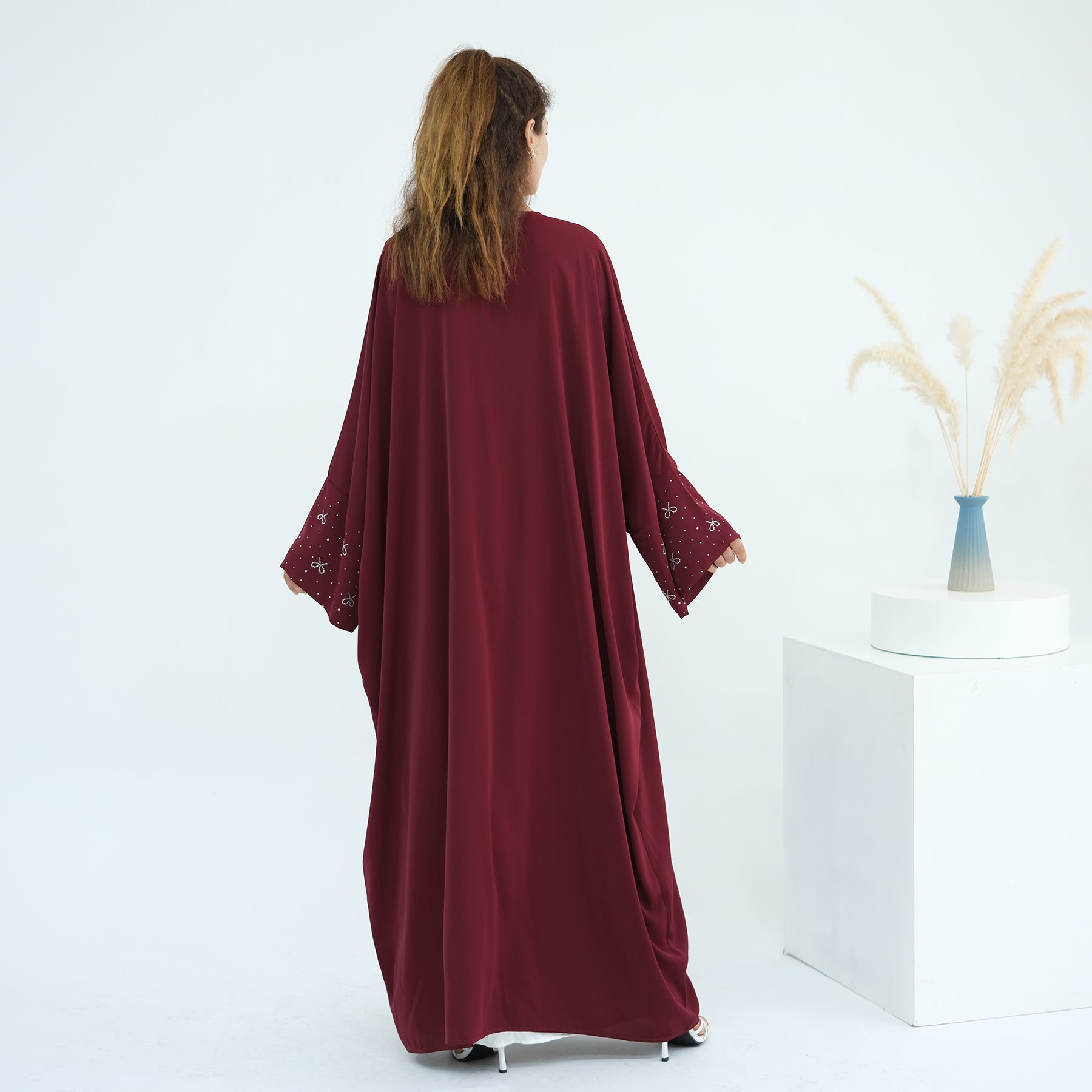 Woman wearing a burgundy abaya with floral lace sleeves on a white background. AlLayaliEmbroideredOpenAbaya_Noor_GraceRed1