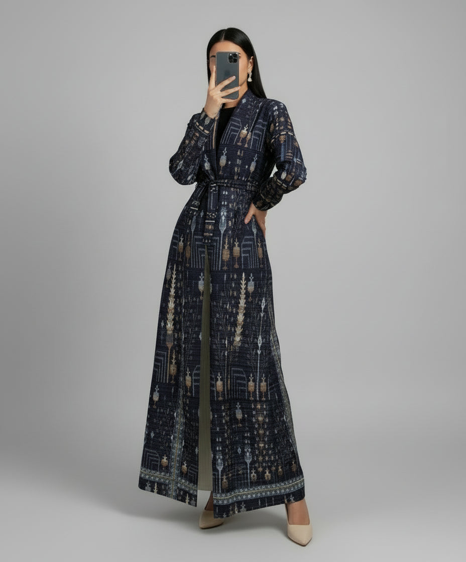 Person wearing a long, patterned dress standing indoors with a plant and chair in the background. Al_Noor_Pleated_Batwing_Abaya_2025_Dubai_Edition_Blue_1 - Noor  & Grace