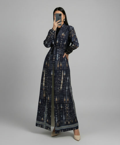 Person wearing a long, patterned dress standing indoors with a plant and chair in the background. Al_Noor_Pleated_Batwing_Abaya_2025_Dubai_Edition_Blue_1 - Noor  & Grace