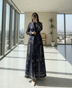 Person wearing a long, patterned dress standing indoors with a plant and chair in the background. Al_Noor_Pleated_Batwing_Abaya_2025_Dubai_Edition_Blue_1_c67328b5-800f-4fd7-873a-9cbe7f35a795 - Noor & Grace