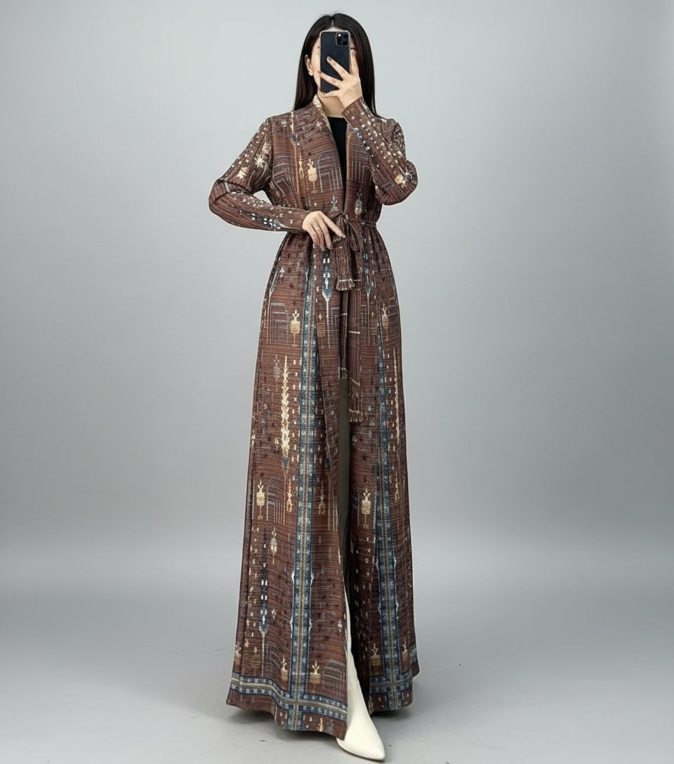 Person wearing a long, patterned coat in an indoor setting with a chair and plant. Al_Noor_Pleated_Batwing_Abaya_2025_Dubai_Edition_Brown_1 - Noor & Grace