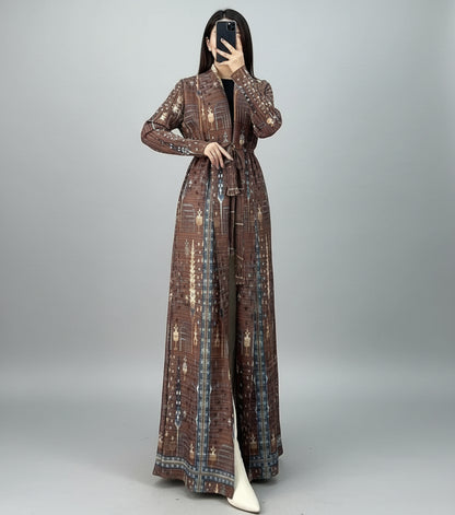 Person wearing a long, patterned coat in an indoor setting with a chair and plant. Al_Noor_Pleated_Batwing_Abaya_2025_Dubai_Edition_Brown_1 - Noor & Grace