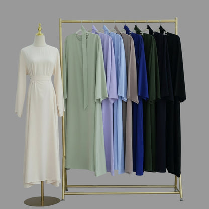 Mannequin with a beige dress and a rack of colorful dresses against a white wall. AsasAbayaInnerDress_Noor_Grace1 - Noor & Grace