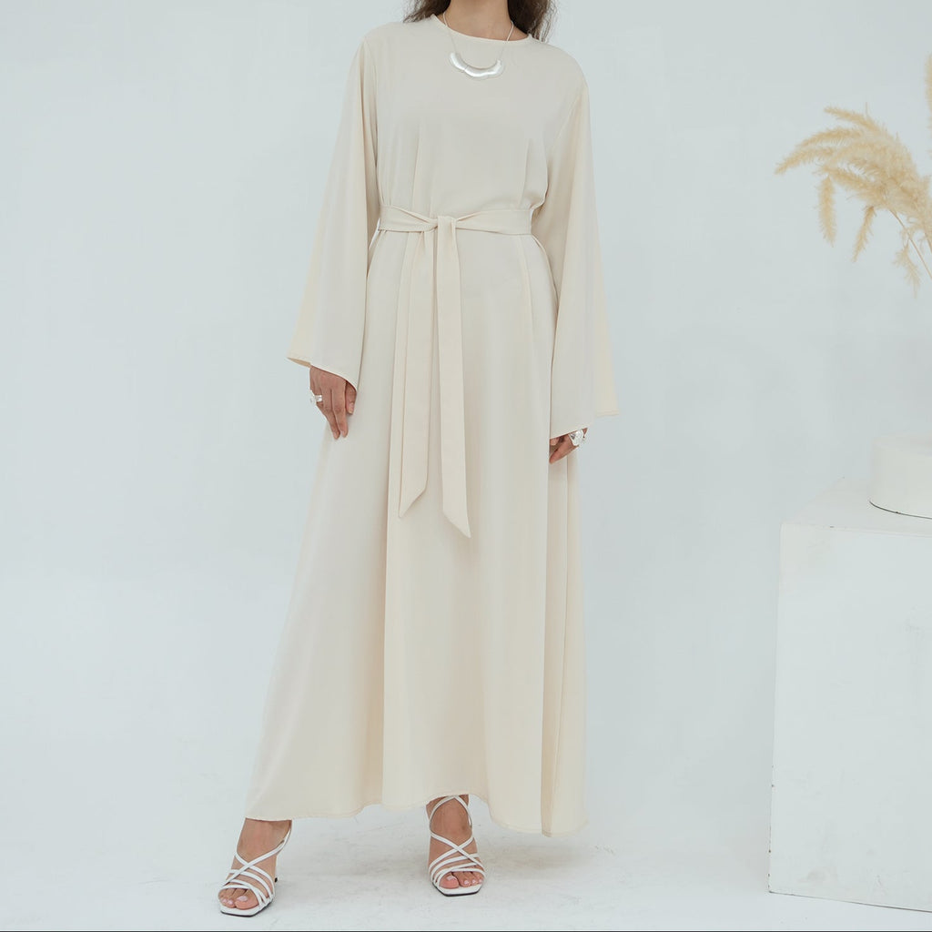 Woman wearing a long beige dress standing against a white background with a vase of dried plants.