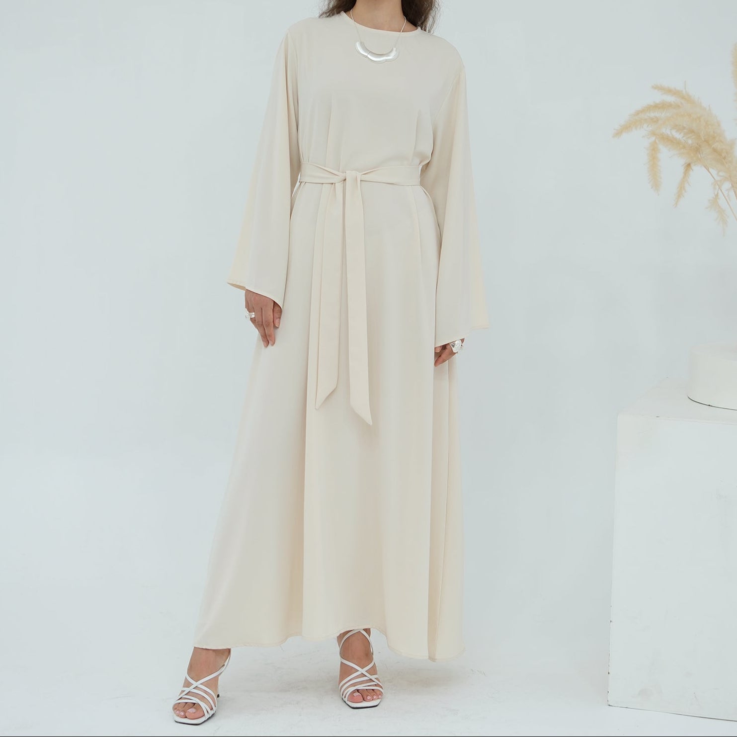 Woman wearing a long beige dress standing against a white background with a vase of dried plants.