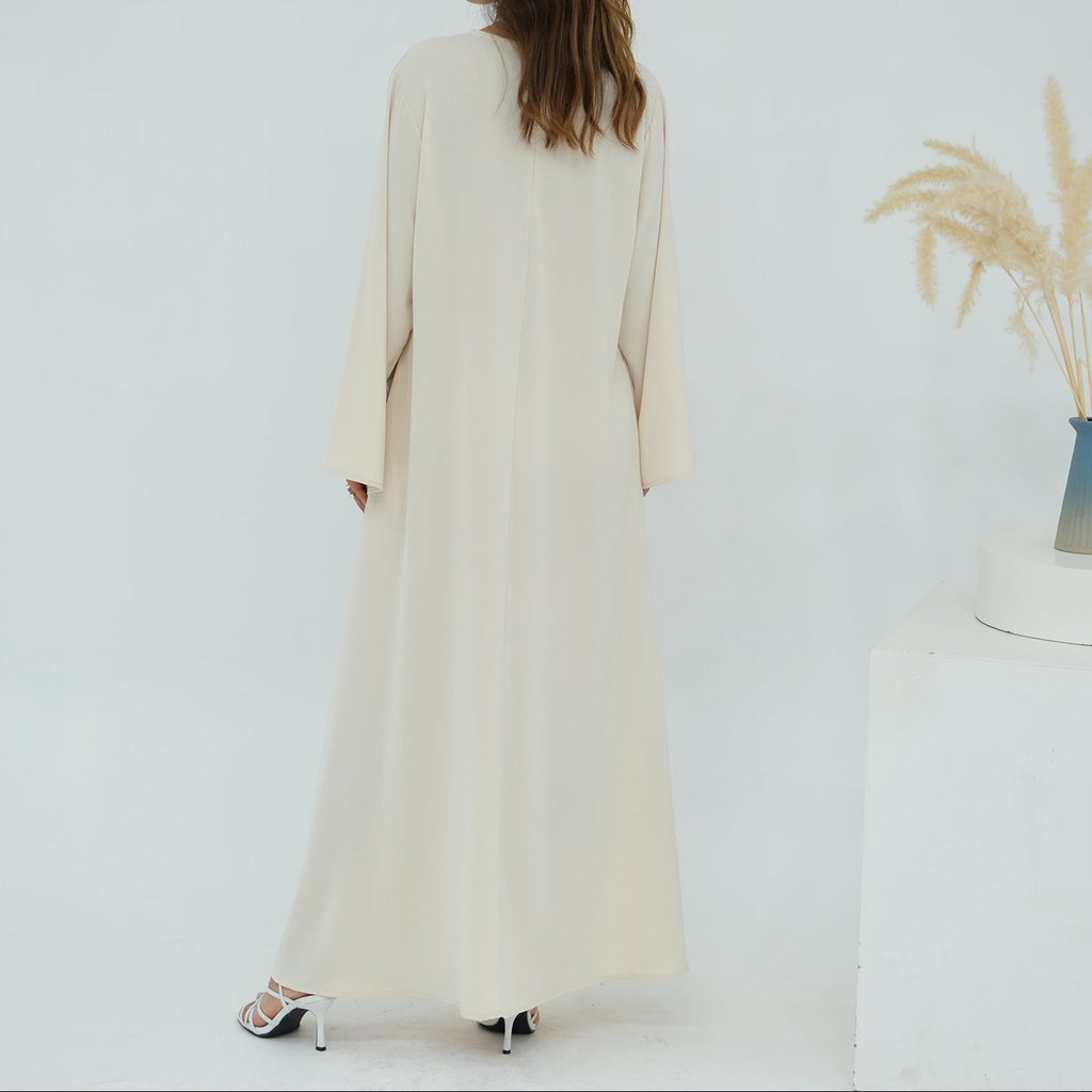 Woman wearing a long cream dress standing in a minimalistic room with a vase of dried plants on a table.