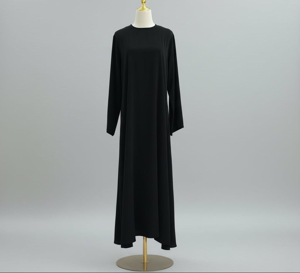 Black dress on a mannequin against a white paneled wall. AsasAbayaInnerDress_Noor_GraceBlack2 - Noor & Grace