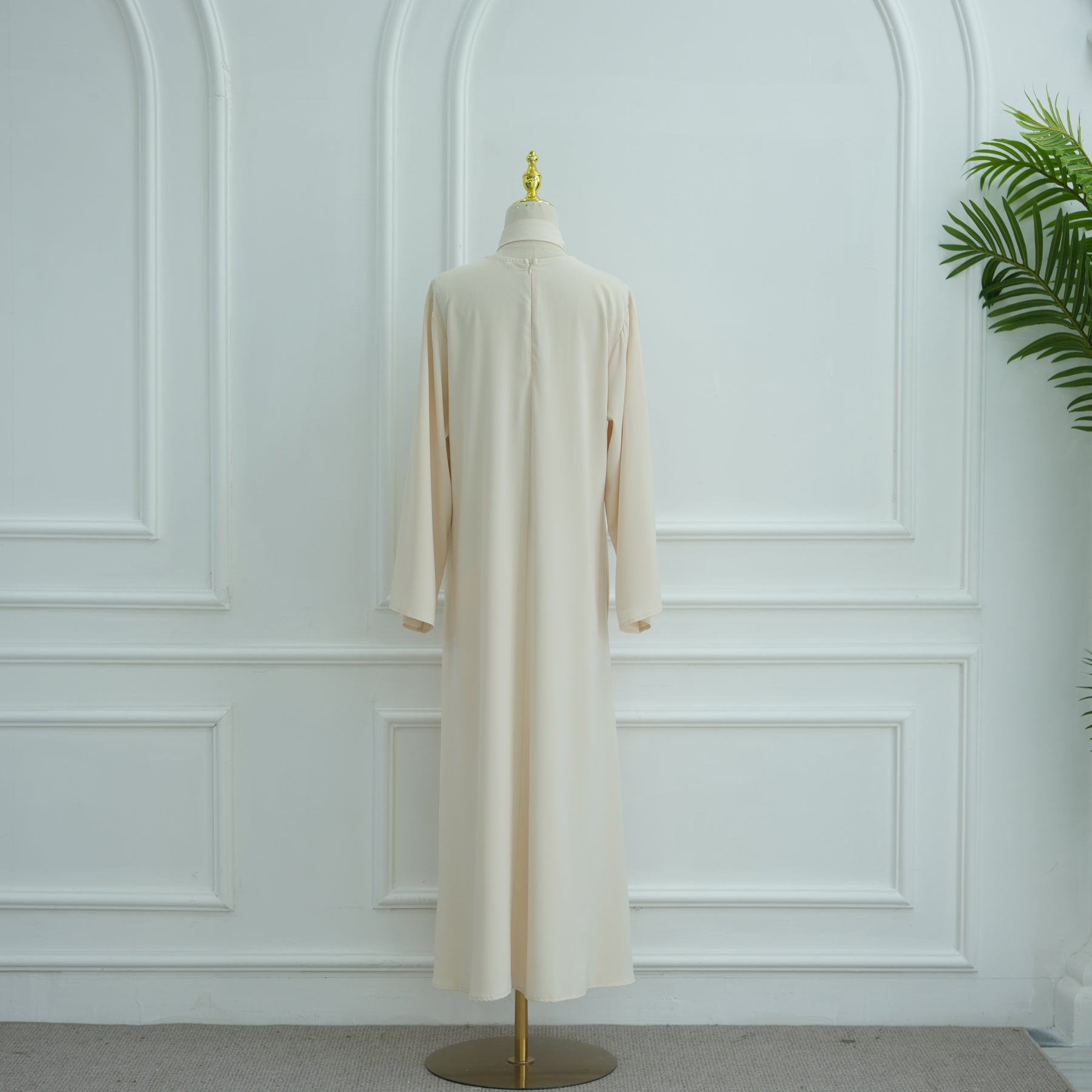 Beige dress on a mannequin against a white paneled wall with a plant in the corner. AsasAbayaInnerDress_Noor_Grace Beige - Noor & Grace