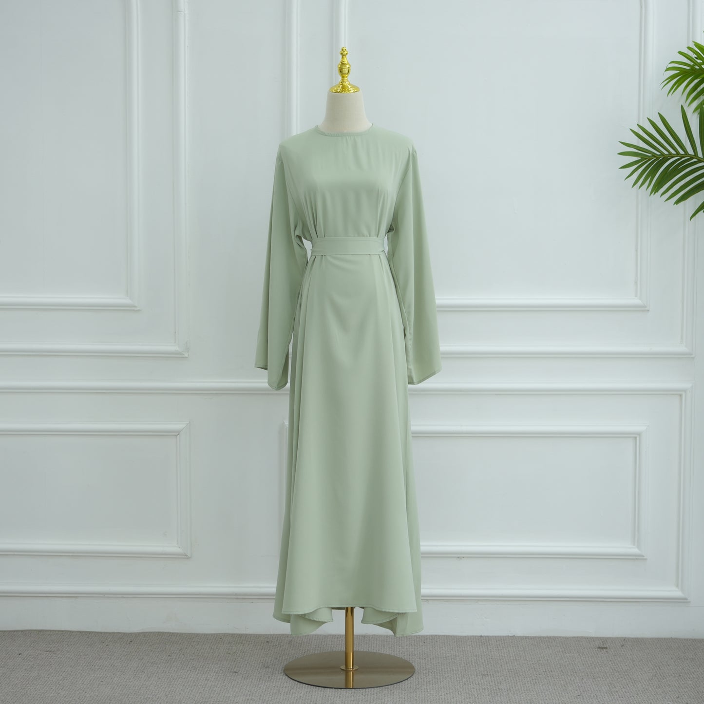 Green dress on a mannequin against a white paneled wall. AsasAbayaInnerDress_Noor_GraceGreen - Noor & Grace