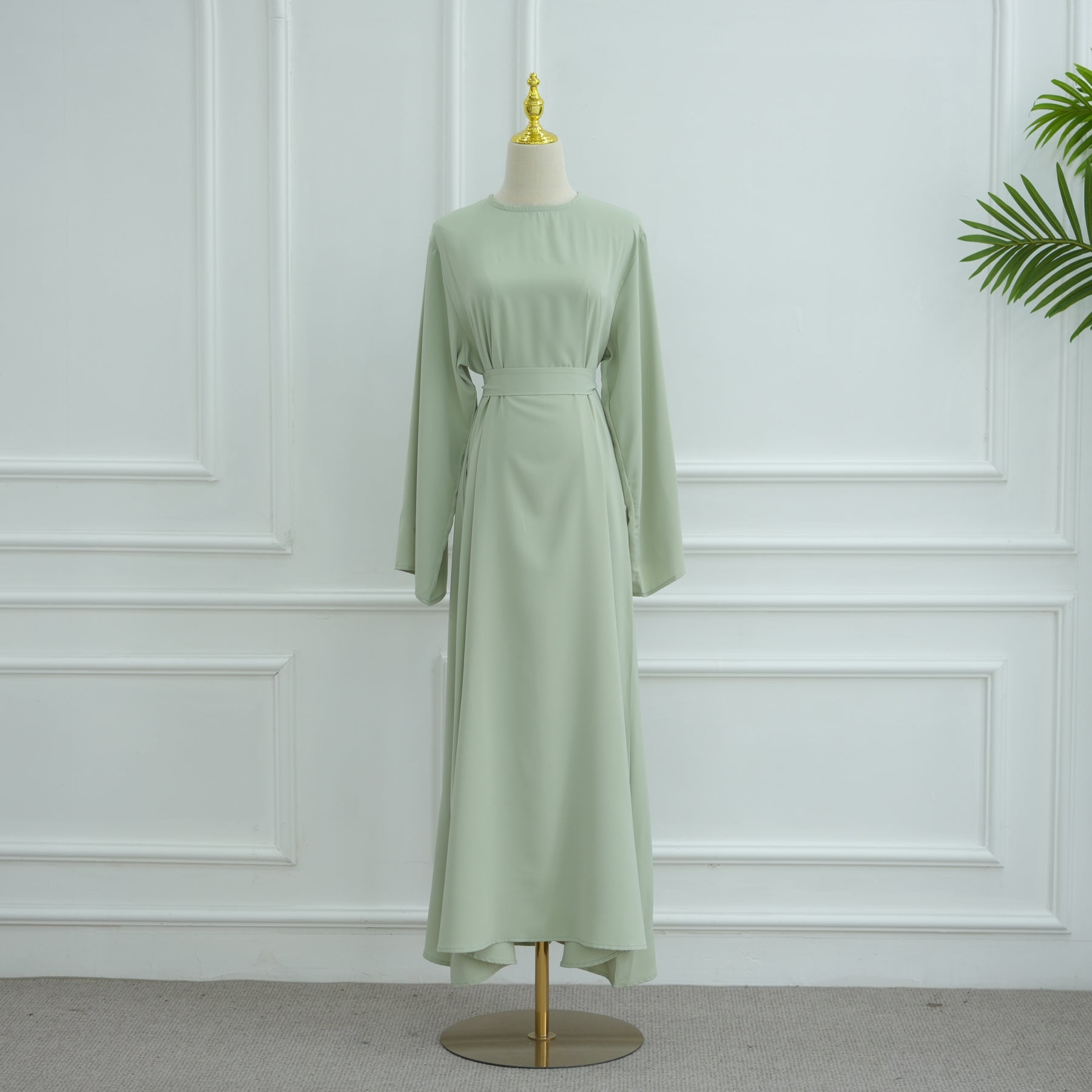 Green dress on a mannequin against a white paneled wall. AsasAbayaInnerDress_Noor_GraceGreen - Noor & Grace