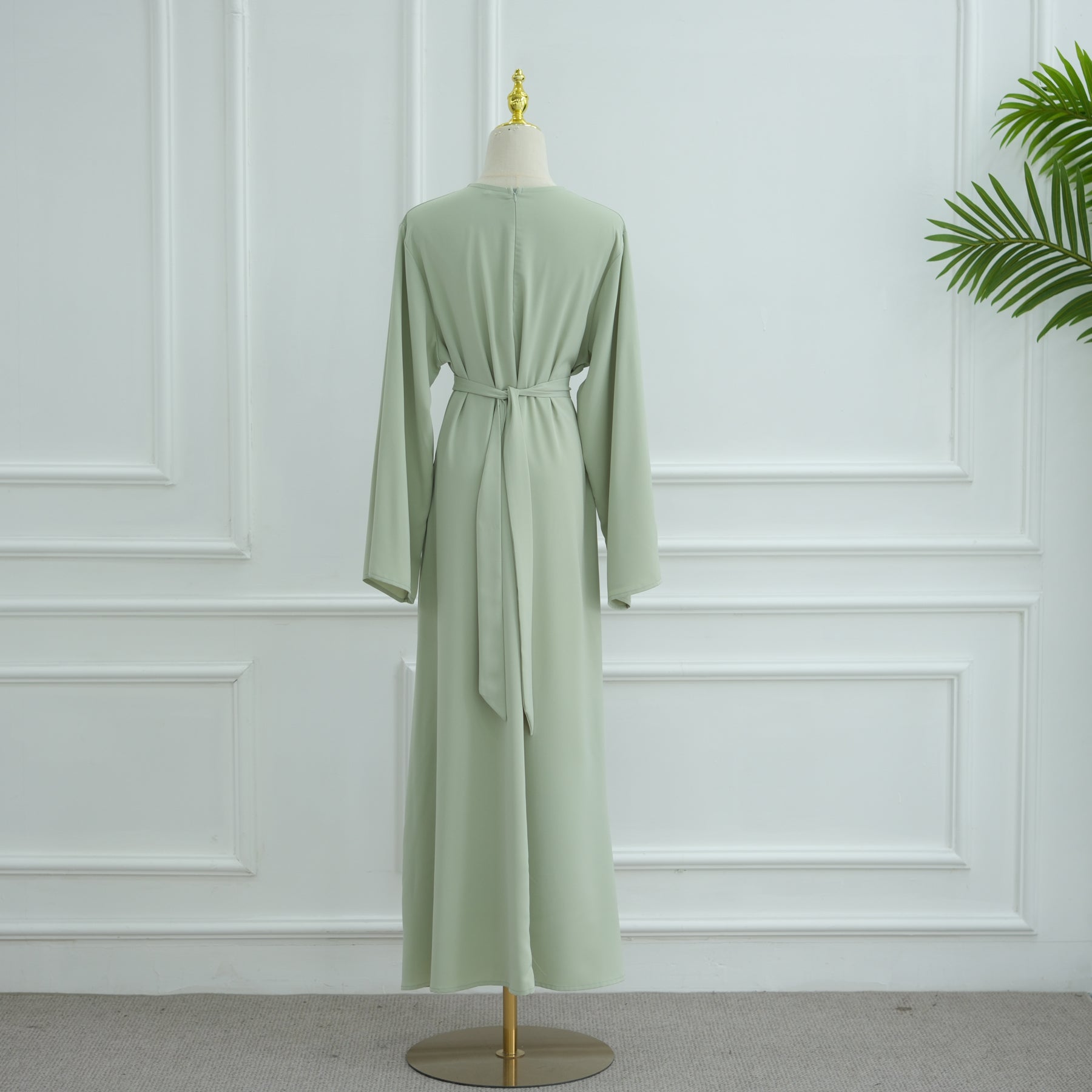 Green dress on a mannequin against a white paneled wall. AsasAbayaInnerDress_Noor_GraceGreen - Noor & Grace