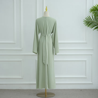 Green dress on a mannequin against a white paneled wall. AsasAbayaInnerDress_Noor_GraceGreen - Noor & Grace