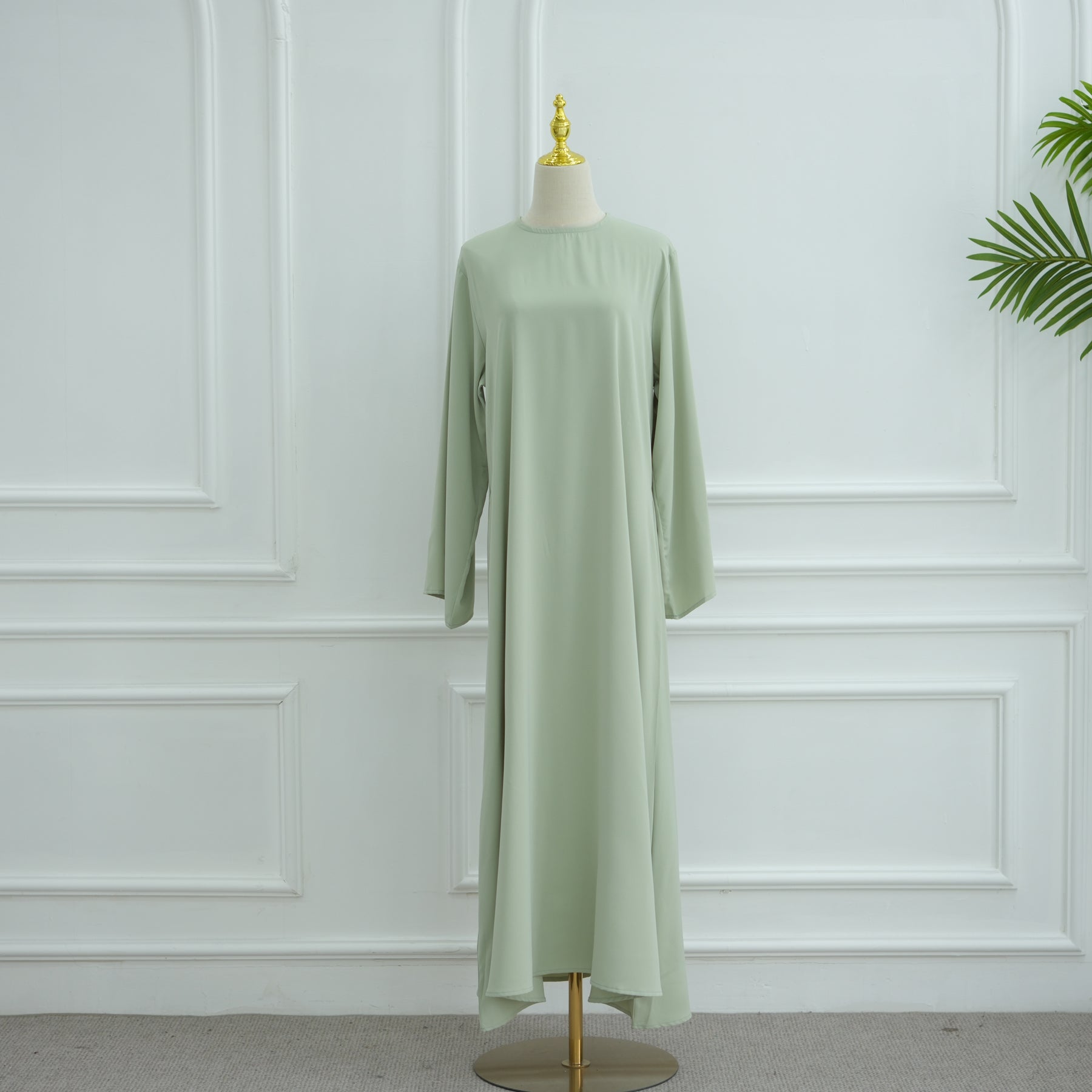 Light green dress on a mannequin against a white paneled wall. AsasAbayaInnerDress_Noor_GraceGreen - Noor & Grace