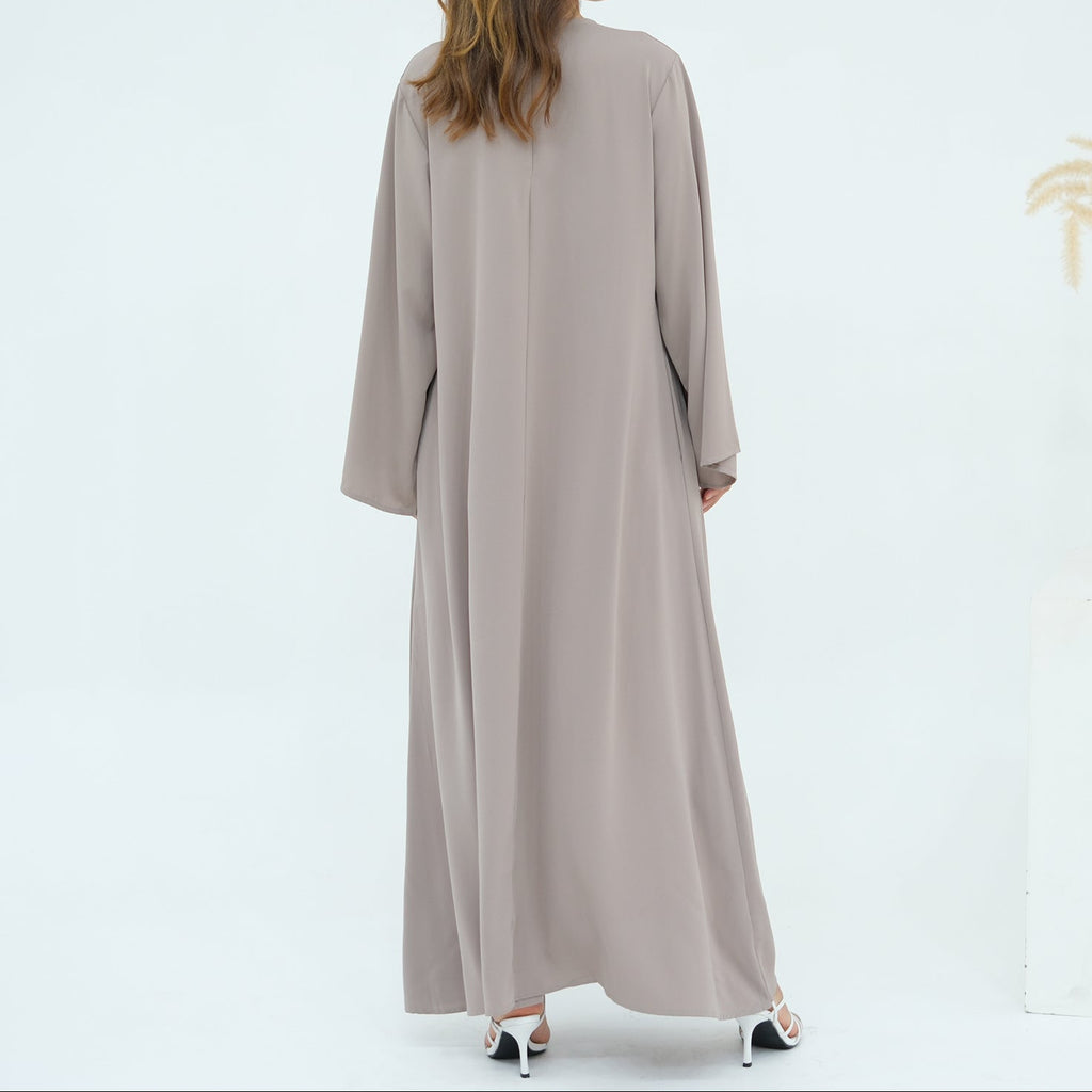 Woman wearing a long beige dress standing in front of a white wall with a vase of dried plants.