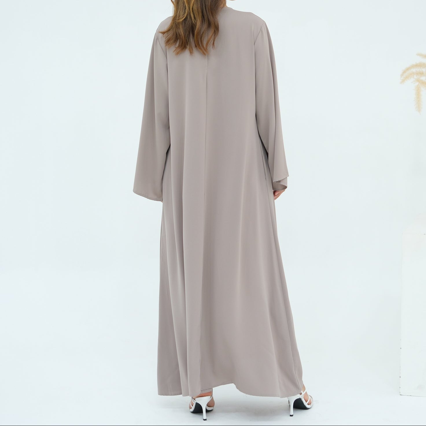 Woman wearing a long beige dress standing in front of a white wall with a vase of dried plants.