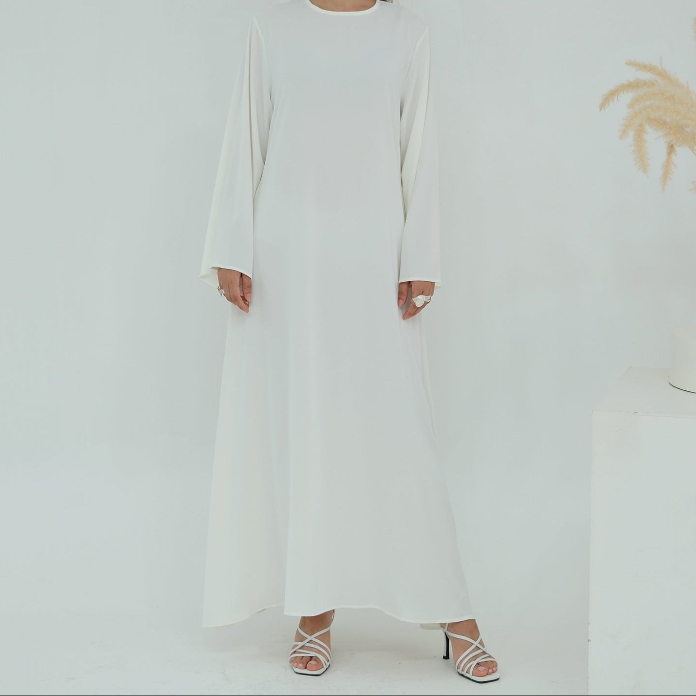 Woman wearing a long white dress standing against a plain background with a vase of dried plants.