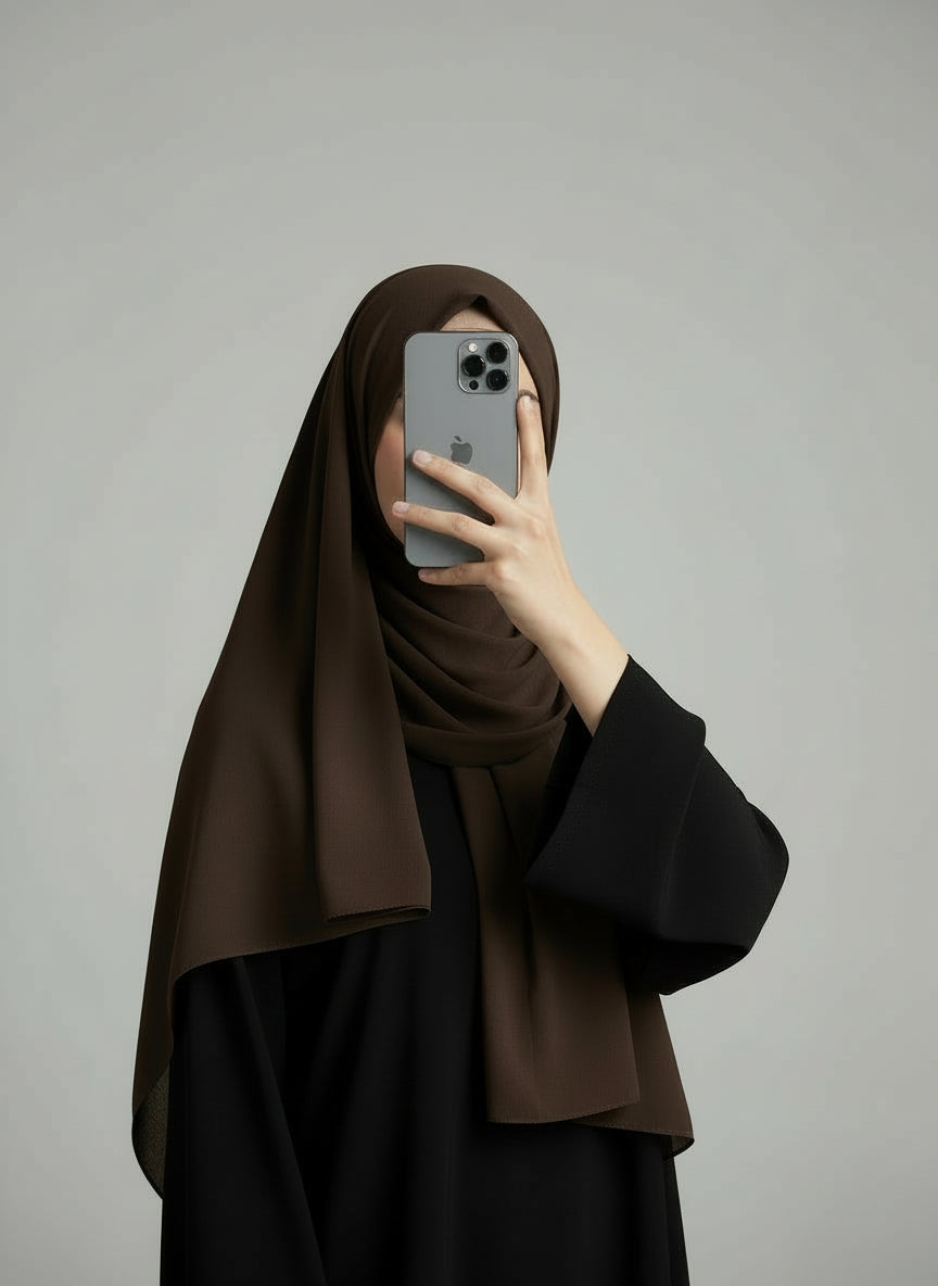 Person wearing a brown hijab and black outfit holding a phone on a gray background. Coffee_Chiffon_Hijab Noor & Grace