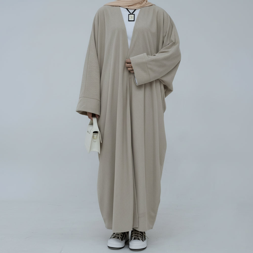 Person wearing a beige corduroy abaya with a white background and a vase with dried plants on the left. EverydayCorduroyAbaya_Beige1 - Noor & Grace