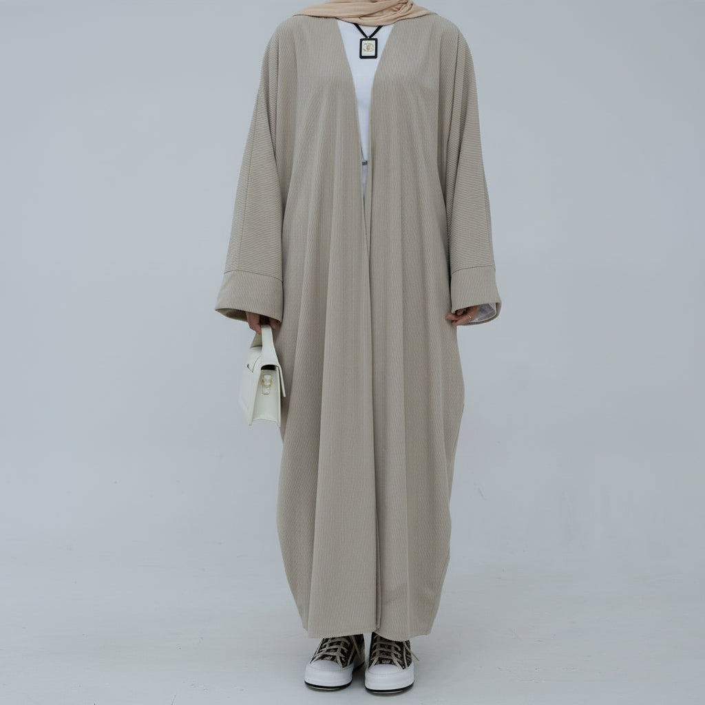 Person wearing a beige corduroy abaya with a white background and a vase with dried plants on the left. EverydayCorduroyAbaya_Beige1 - Noor & Grace