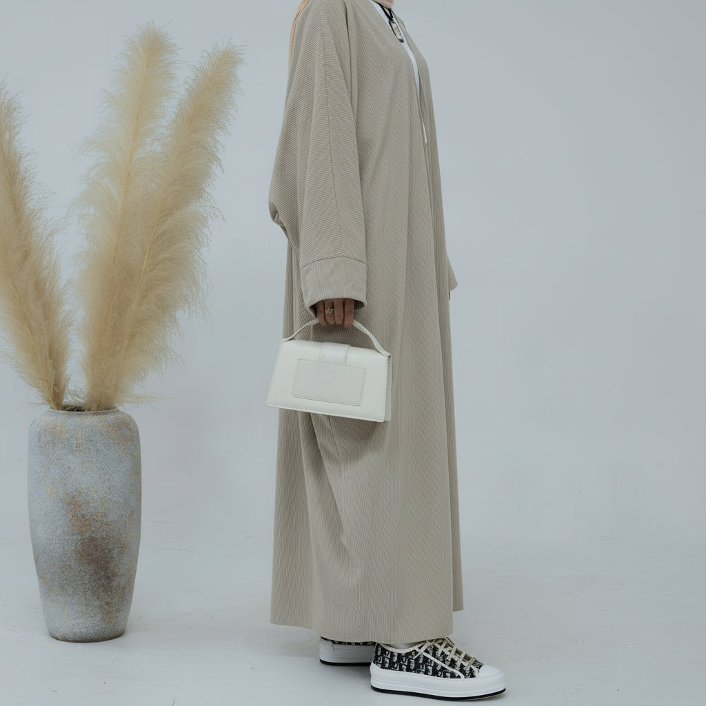 Person wearing a beige corduroy abaya with a white background and a vase with dried plants on the left. EverydayCorduroyAbaya_Beige1 - Noor & Grace