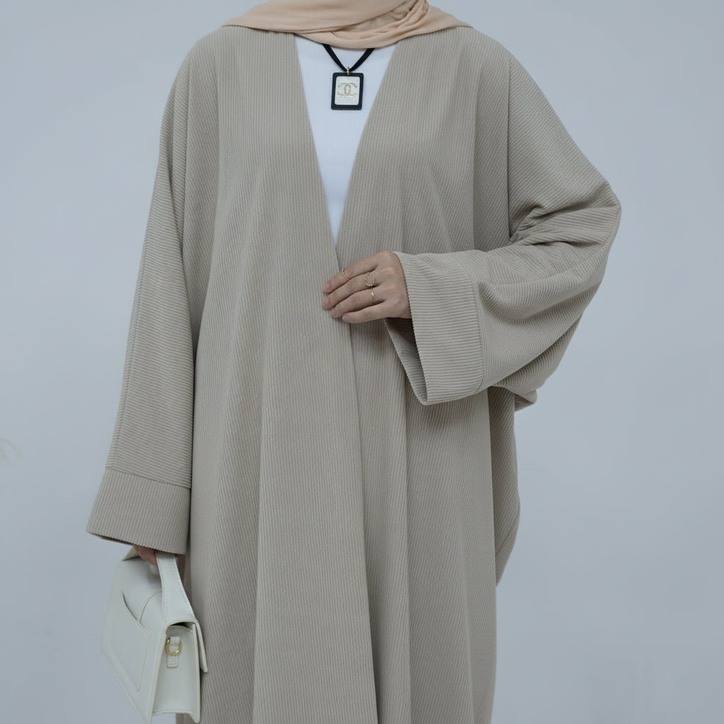 Person wearing a beige corduroy abaya with a white background and a vase with dried plants on the left. EverydayCorduroyAbaya_Beige1 - Noor & Grace