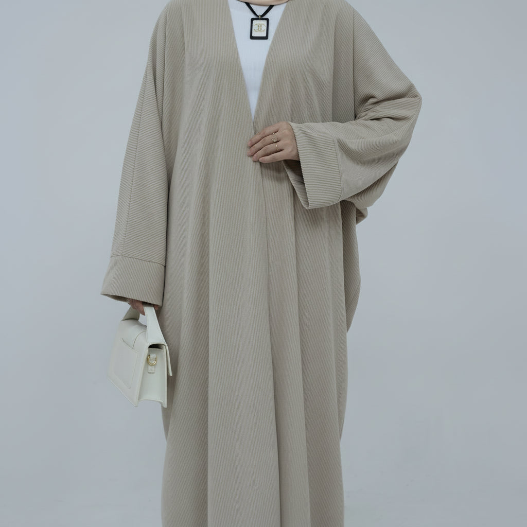 Person wearing a beige corduroy abaya with a white background and a vase with dried plants on the left. EverydayCorduroyAbaya_Beige1 - Noor & Grace