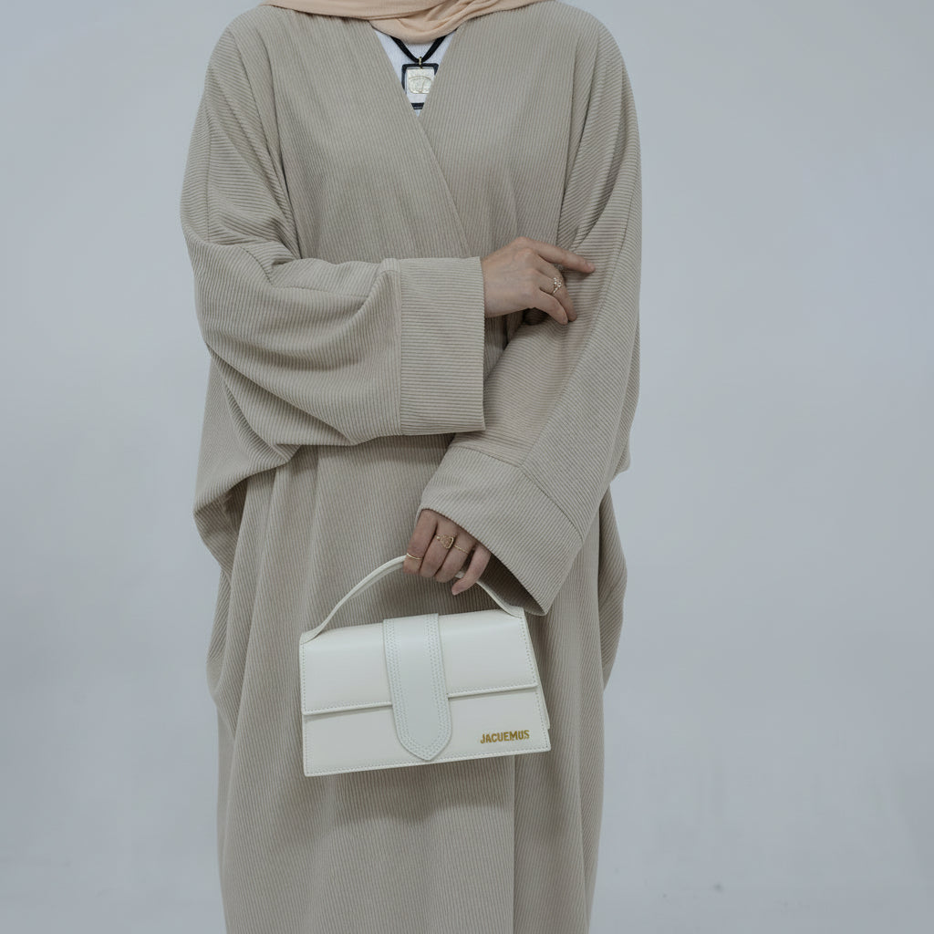 Person wearing a beige corduroy abaya with a white background and a vase with dried plants on the left. EverydayCorduroyAbaya_Beige1 - Noor & Grace