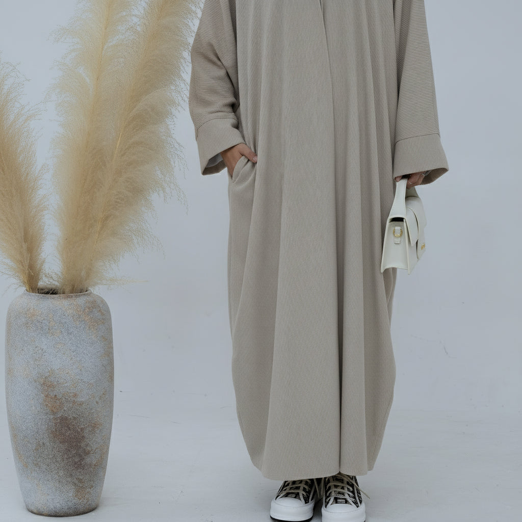 Person wearing a beige corduroy abaya with a white background and a vase with dried plants on the left. EverydayCorduroyAbaya_Beige1 - Noor & Grace