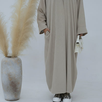 Person wearing a beige corduroy abaya with a white background and a vase with dried plants on the left. EverydayCorduroyAbaya_Beige1 - Noor & Grace