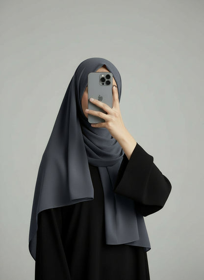 Person wearing a black abaya and gray hijab holding a phone on a plain background. Gray_Chiffon_Hijab Noor & Grace