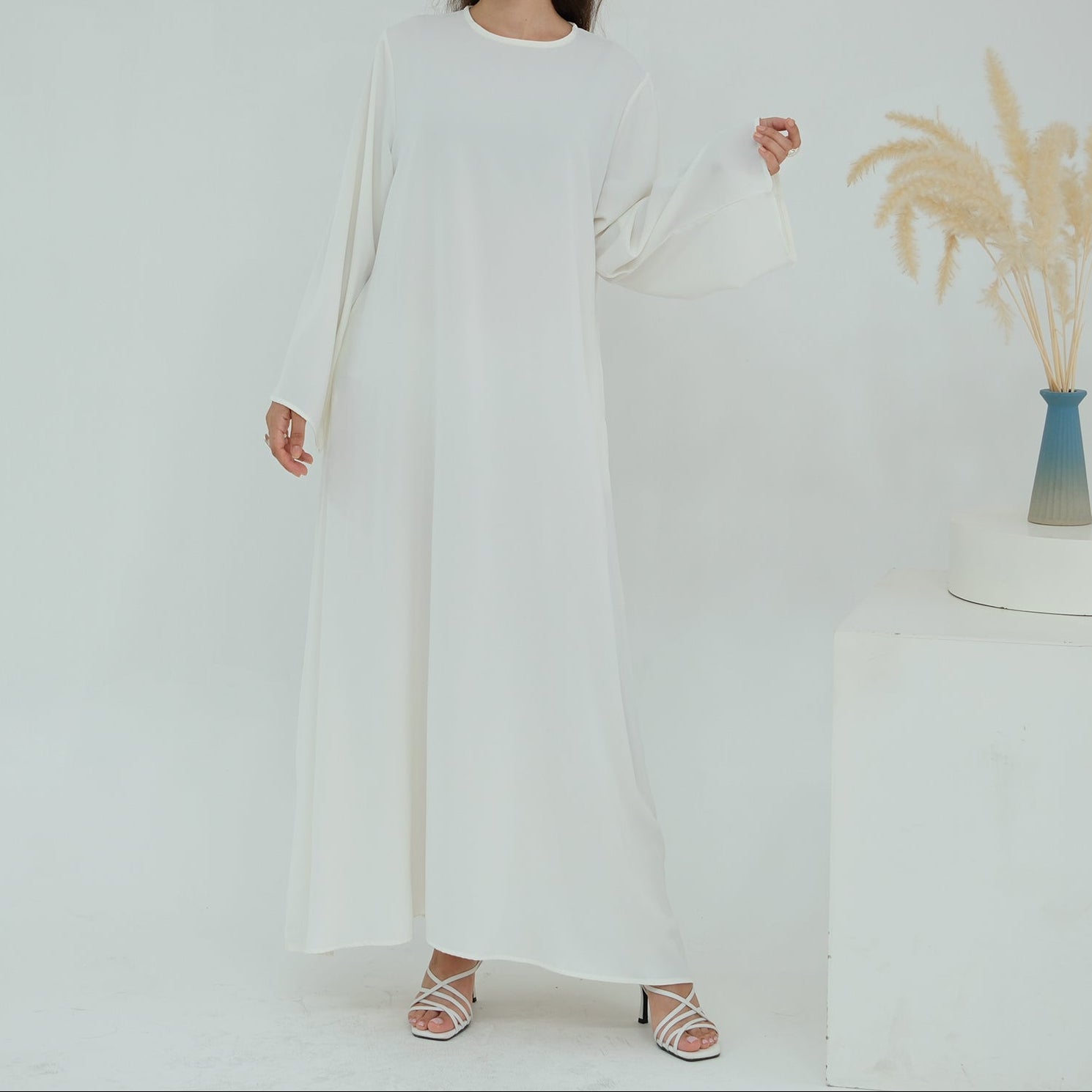 Woman wearing a long white dress standing against a plain background with a vase of dried plants.