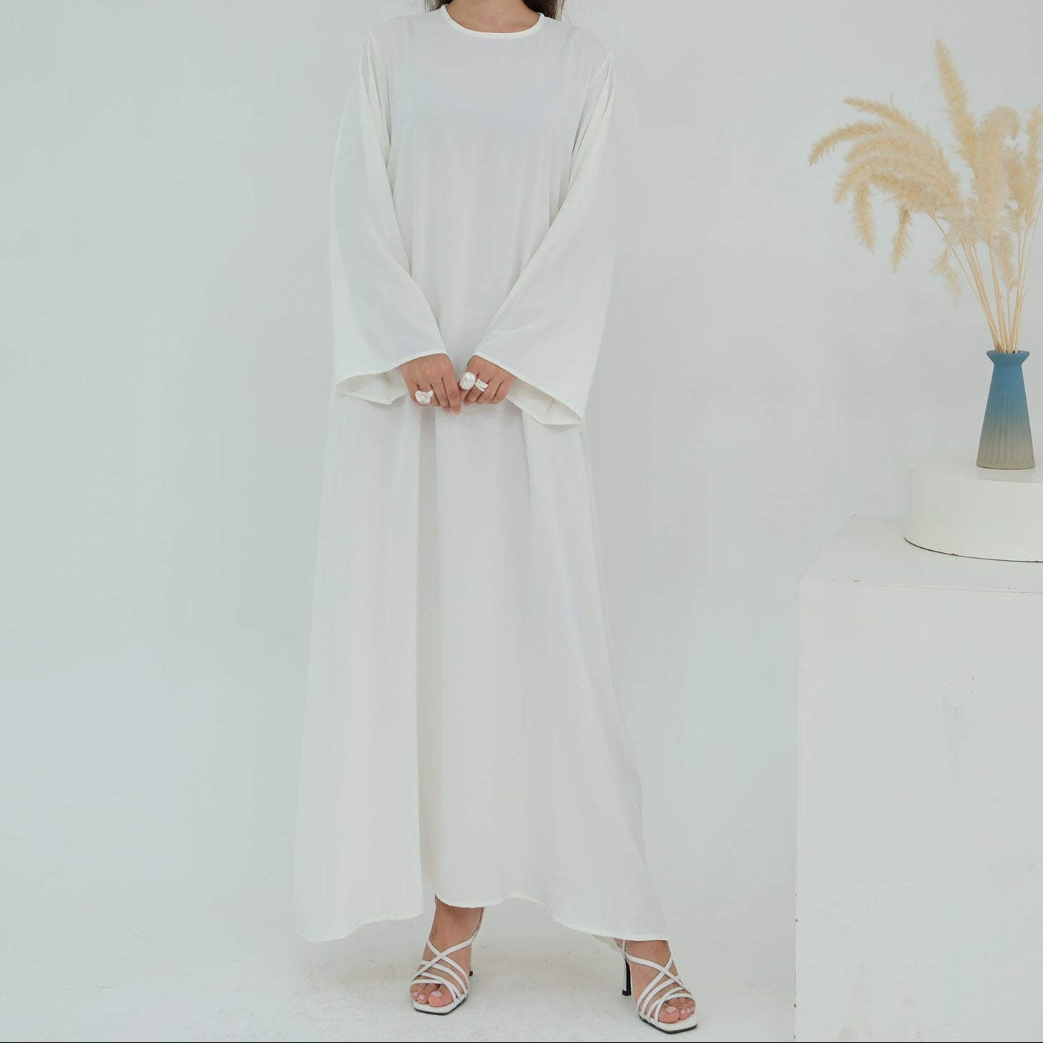 Woman wearing a white outfit standing in a minimalistic room with a vase of dried plants.