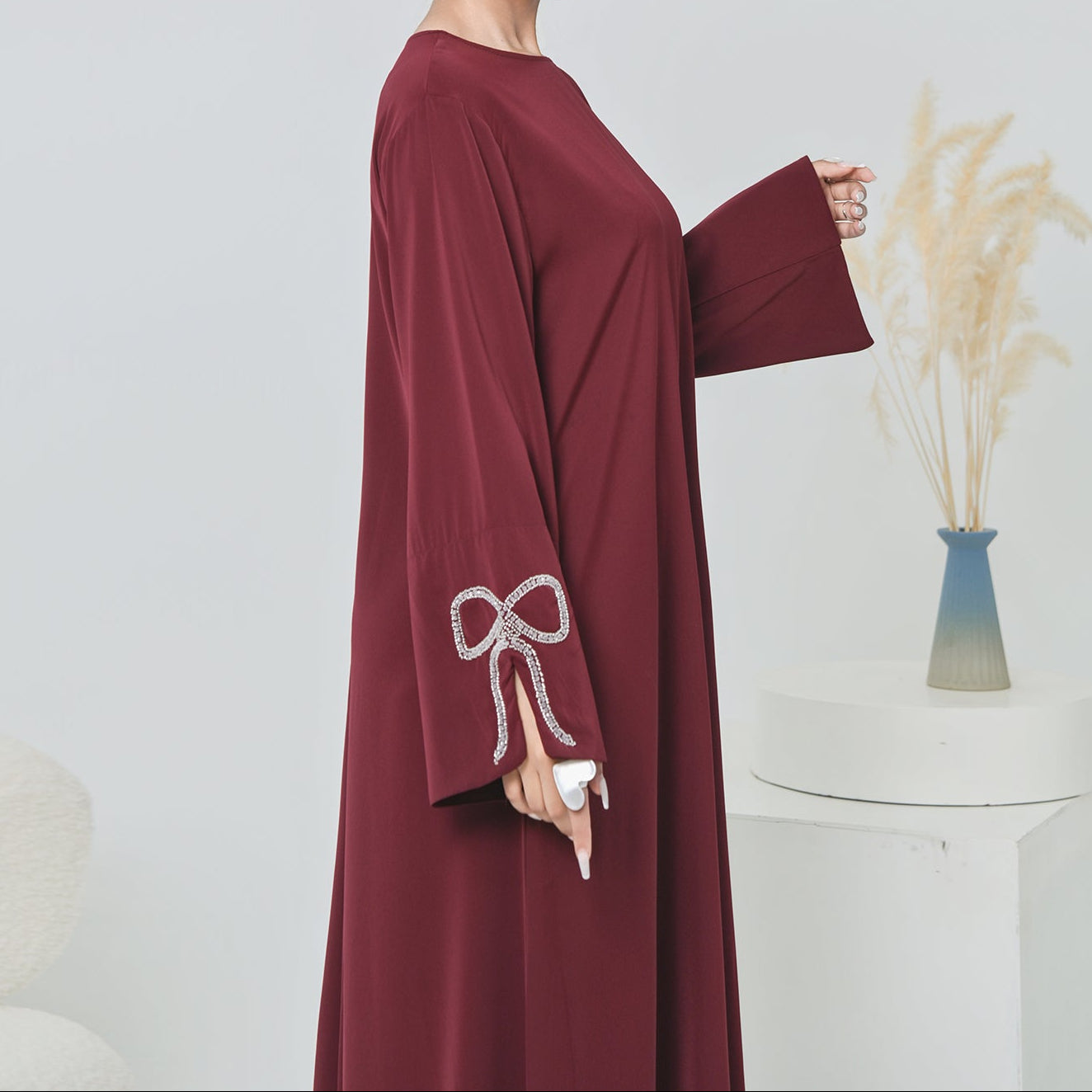 Woman wearing a burgundy abaya with a bow detail, standing against a plain background.