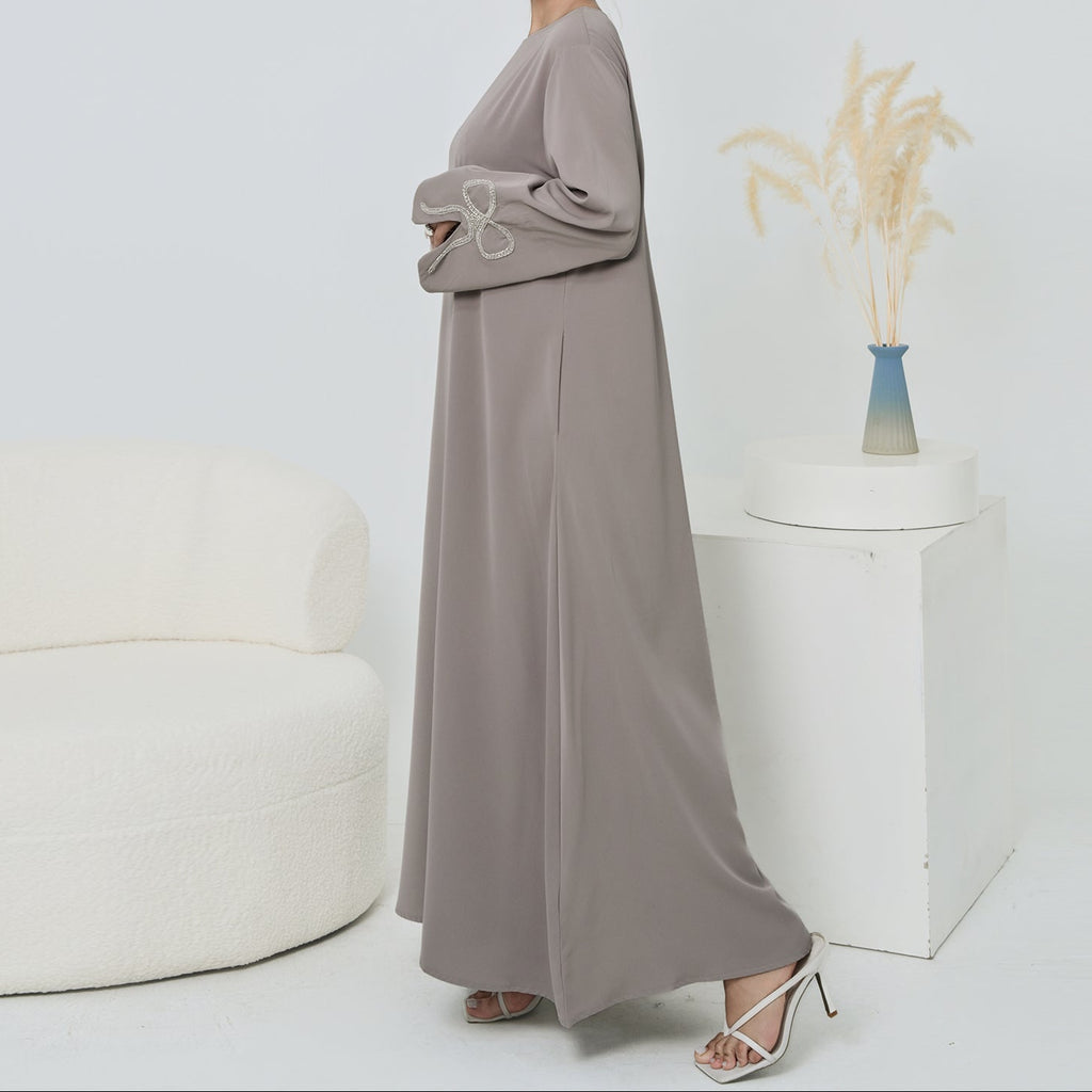 Woman wearing a long, light gray dress standing in a minimalistic room with a white wall and a small decorative plant.