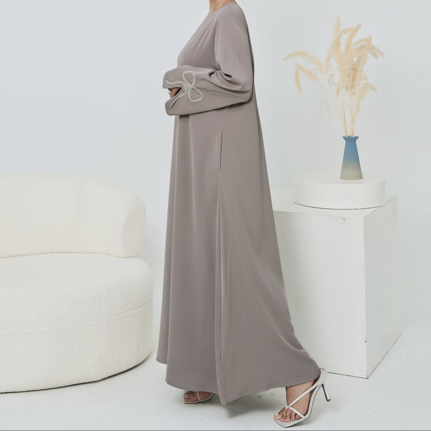 Woman wearing a long, light gray dress standing in a minimalistic room with a white wall and a small decorative plant.