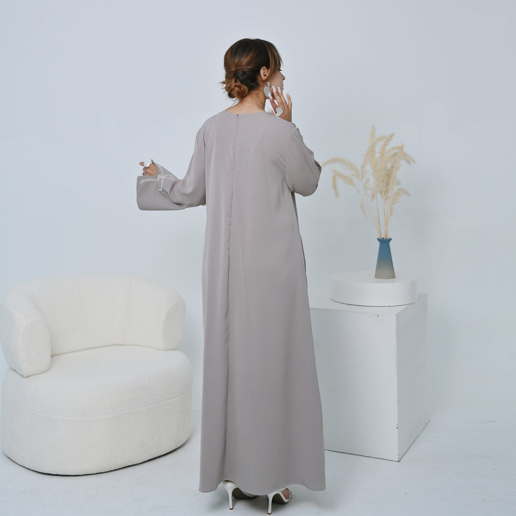 Woman in a long light gray dress standing in a minimalistic room with a white chair and plant.