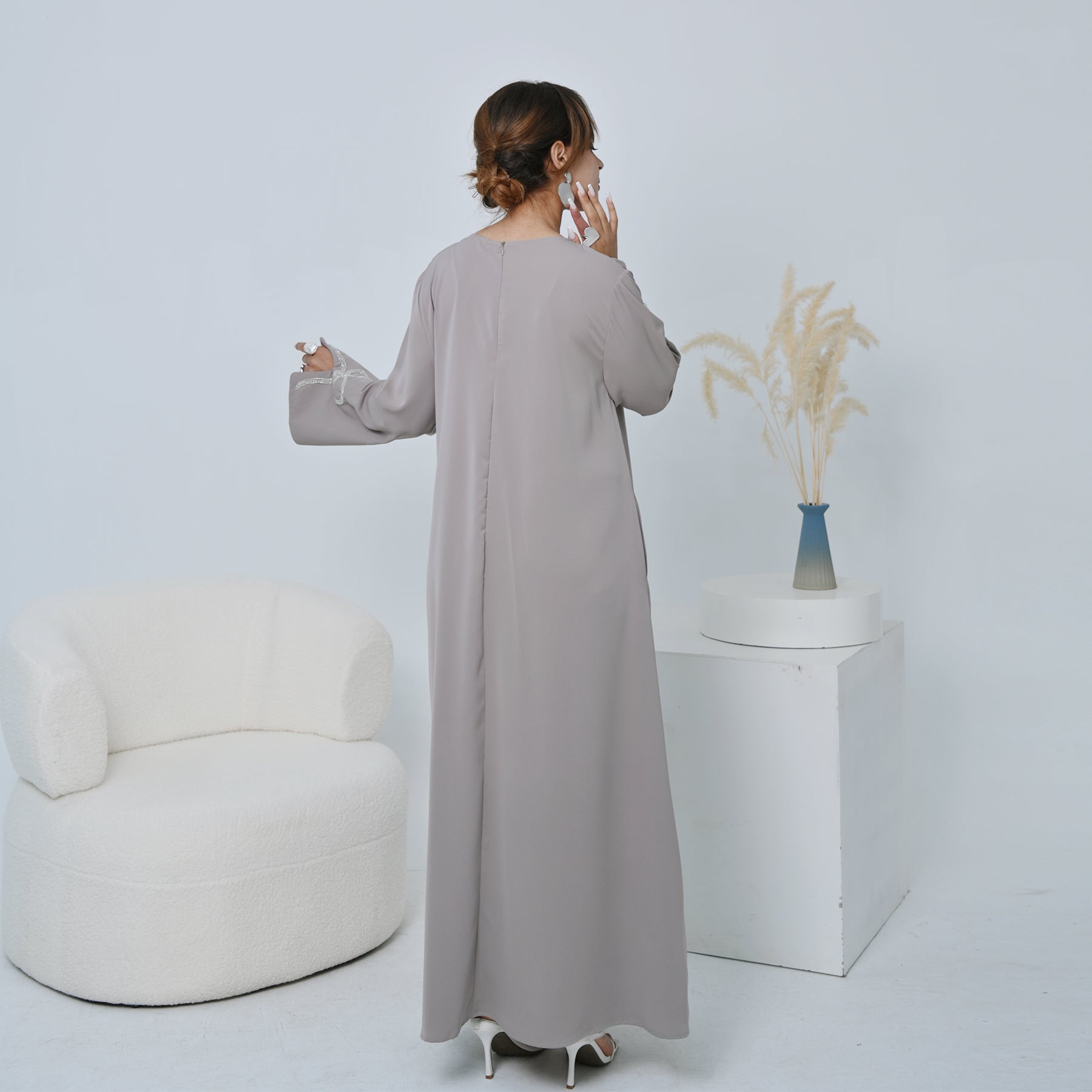 Woman in a long light gray dress standing in a minimalistic room with a white chair and plant.