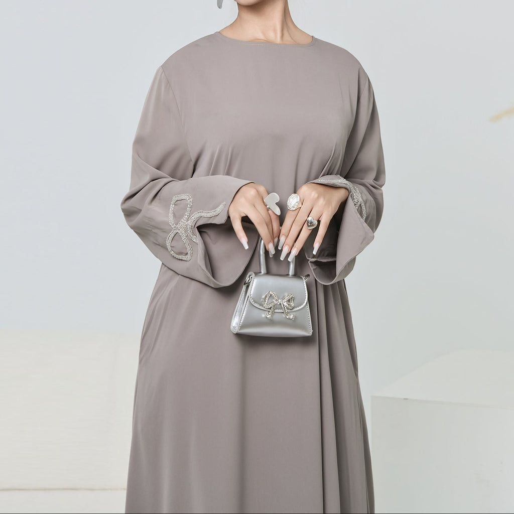 Woman in a long, light gray dress holding a matching handbag against a plain background