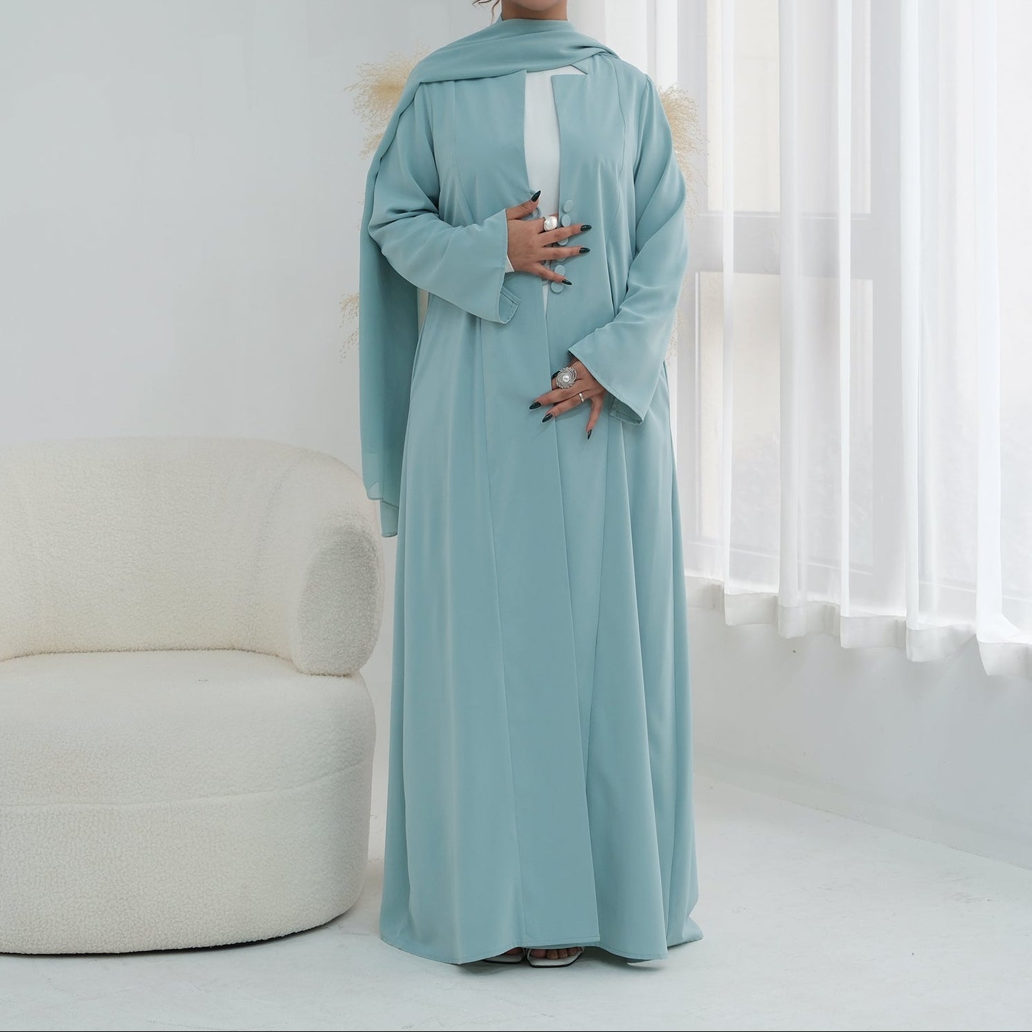 Woman wearing a light blue abaya in a minimalistic room with white furniture and curtains.