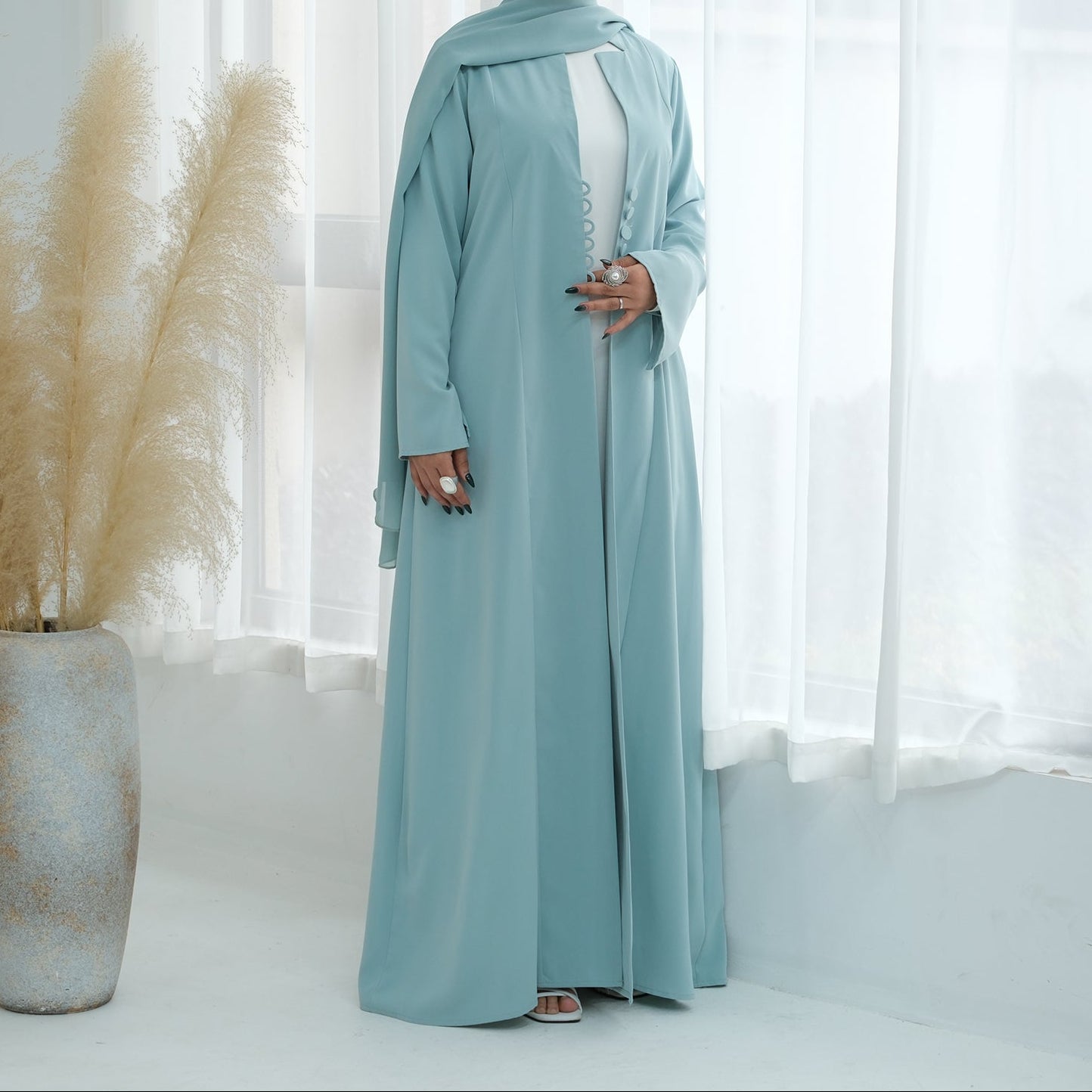 Woman in a light blue abaya standing in a room with white curtains and decorative plants.