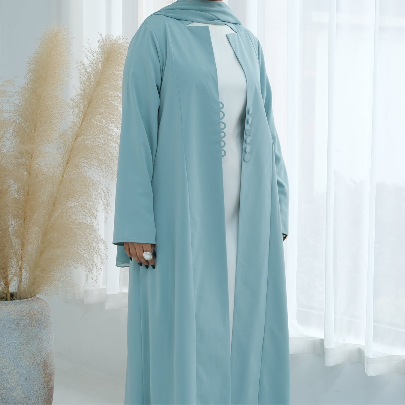 Woman wearing a light blue abaya standing in front of white curtains with decorative plants.