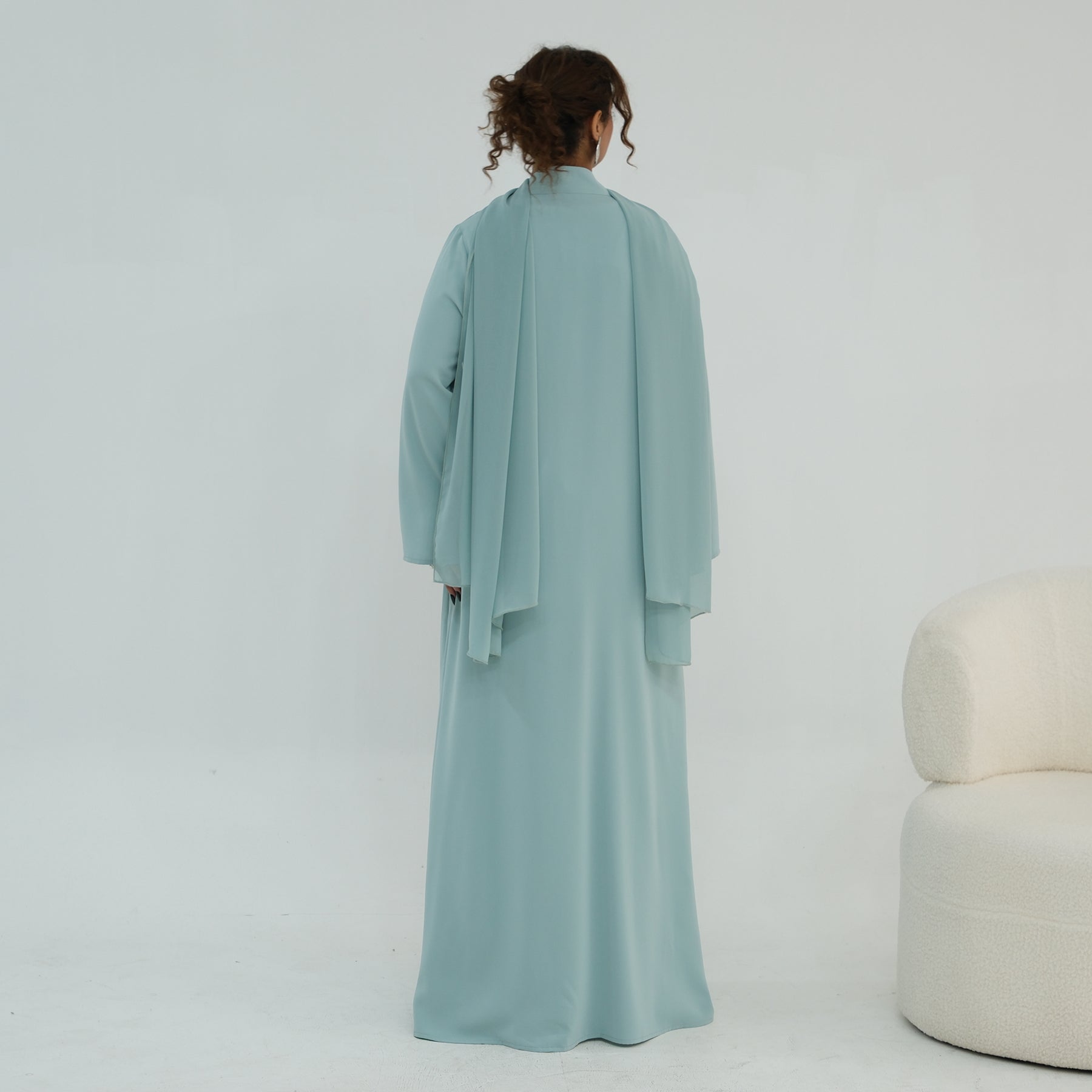 Person wearing a light blue dress standing in a minimalistic room with a white chair.