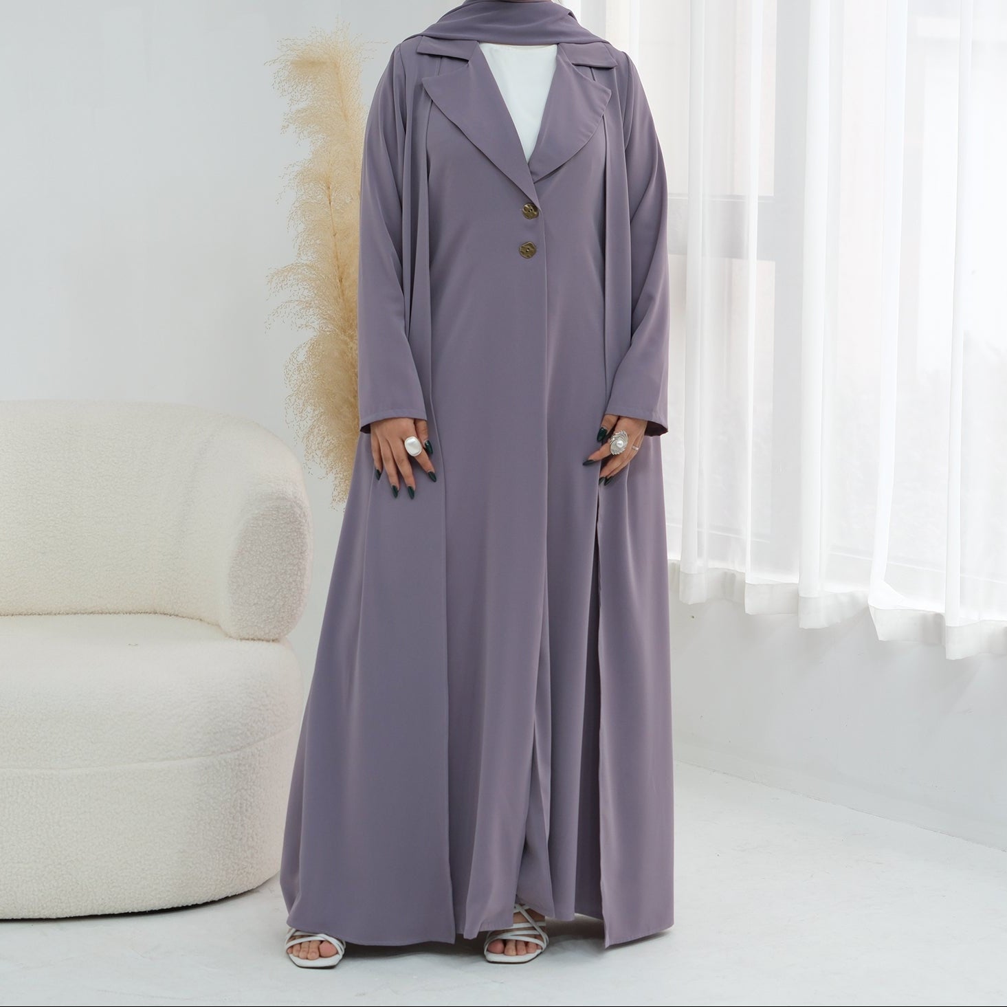 Woman wearing a long, light purple coat in a bright room with white curtains.