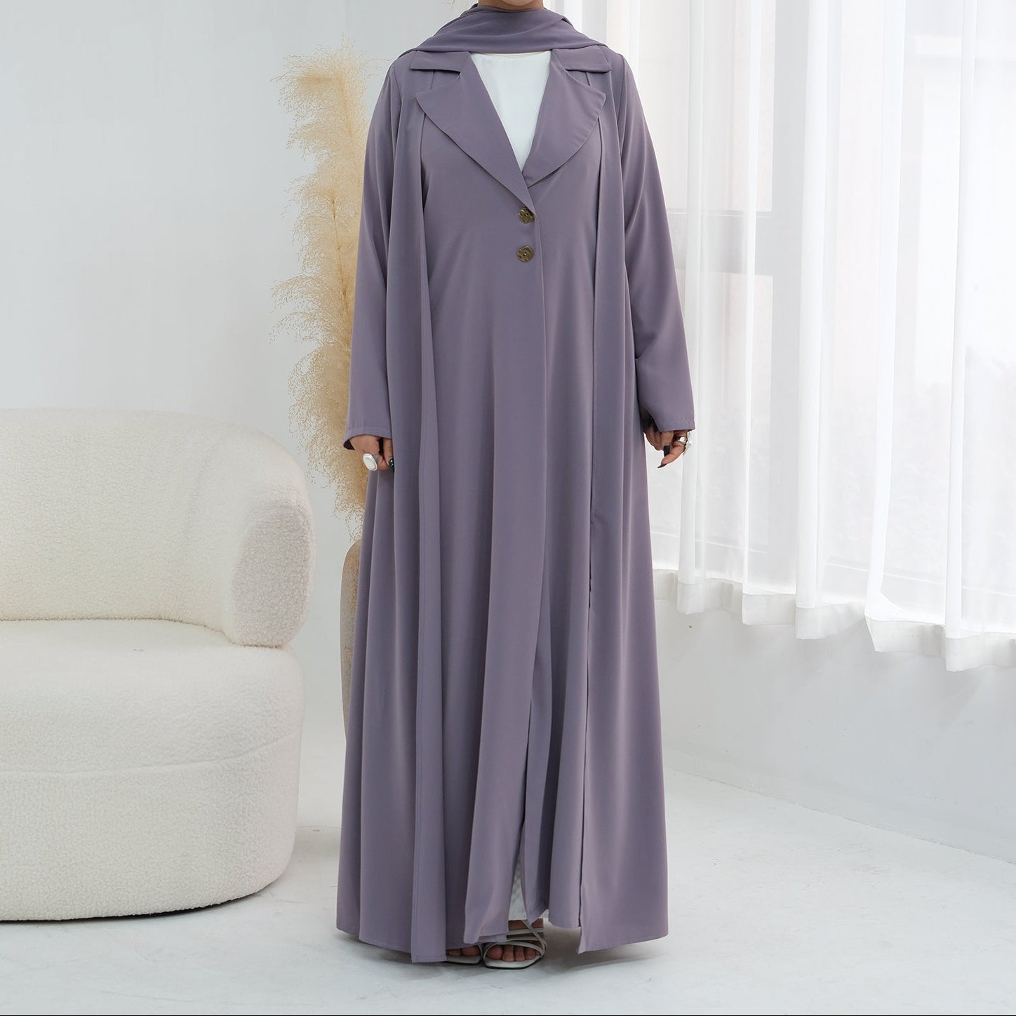 Woman wearing a long, light purple dress with a white top underneath, standing in a bright room.