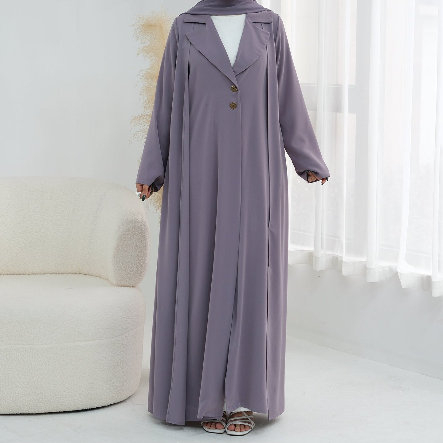 Woman wearing a long, light purple coat in a minimalistic room with white curtains.