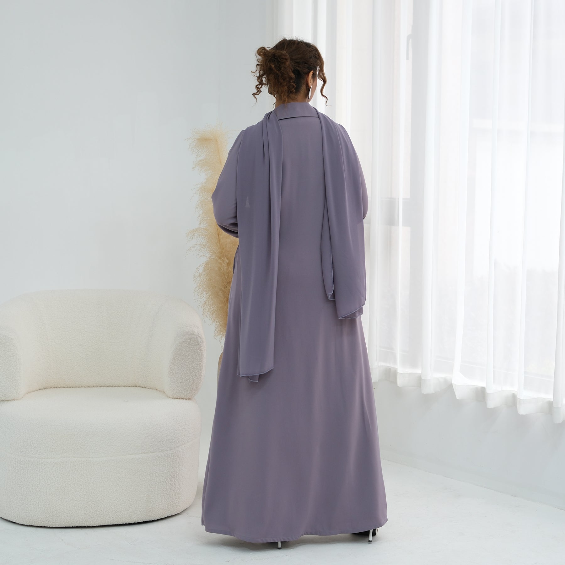 Person wearing a long, light purple dress standing in a minimalistic room with white walls and a white chair.